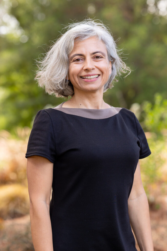 headshot of Milena smiling at the camera, wearing a black top. She stands outside, with a green blurred tree background,
