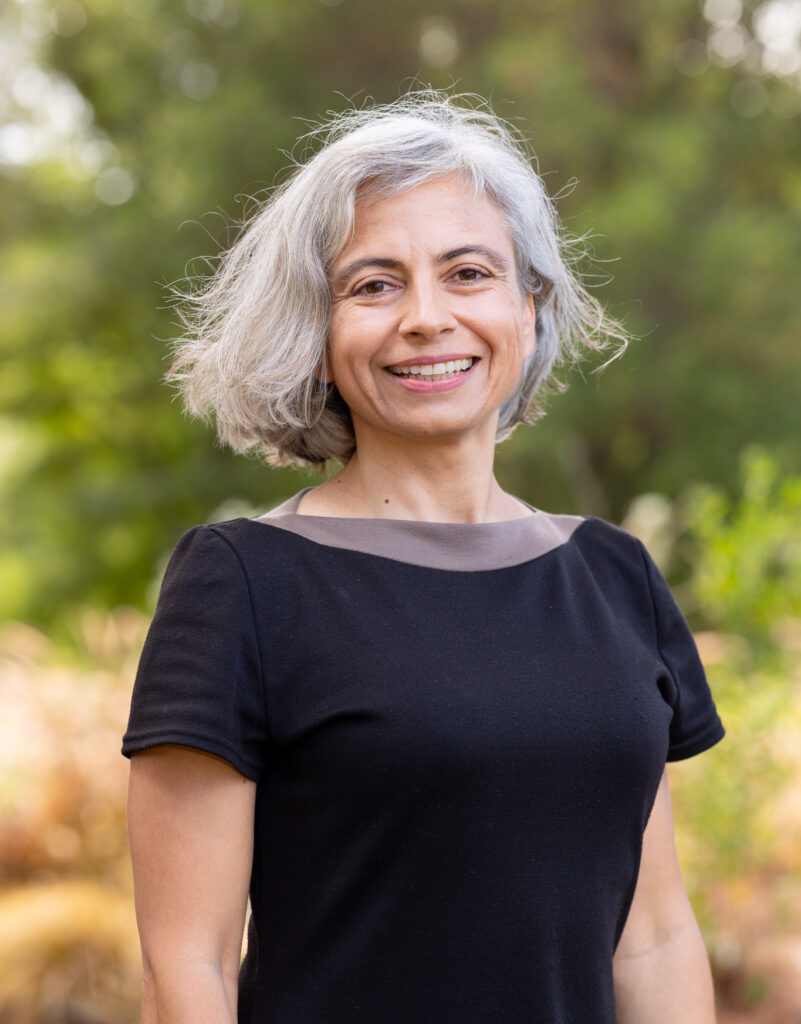 headshot of Milena smiling at the camera, wearing a black top. She stands outside, with a green blurred tree background,